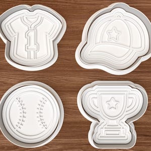 May include: A set of four white plastic cookie cutters featuring sports-themed shapes: a baseball jersey with the number one, a baseball cap with a star, a round baseball with stitched seams, and a trophy cup with a star emblem in the centre.