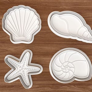 Könnte beinhalten: Vier weiße Ausstecher aus Kunststoff in Form von Muscheln und einem Seestern. Die Ausstecher umfassen eine Jakobsmuschel, eine Muschel, einen Seestern und eine Nautilusmuschel. Die Ausstecher befinden sich auf einer Holzoberfläche.