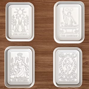 Peut inclure: Quatre emporte-pièces rectangulaires blancs présentant des motifs de cartes de tarot en relief. Les designs incluent The Lovers, The Magician, The Empress et The World, chacun avec son titre respectif imprimé en lettres en relief en bas.