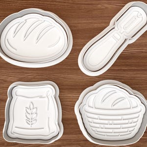 May include: Four white plastic cookie cutters in the shapes of a loaf of bread, a rolling pin, a bag of flour with a wheat stalk, and a bread basket. The cutters are on a brown wooden surface.
