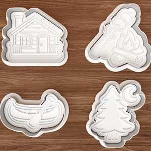May include: Four white plastic cookie cutters in camping-themed shapes. The shapes include a cabin, a campfire with a marshmallow, a canoe with a paddle, and a tree with a crescent moon.