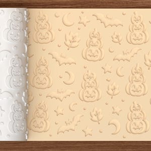 Op de afbeelding: Een witte cilindrische deegroller met een verdiept patroon van gestapelde pompoenen, vleermuizen, maansikkels, sterren en bladeren. Het patroon is in een lichtbeige deeg gedrukt en toont het herhalende ontwerp met een Halloween-thema.