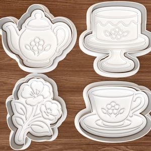 May include: Four white plastic cookie cutters in the shapes of a teapot, a cake on a stand, a floral sprig, and a teacup with saucer. Each cutter has a floral design in the center. The cutters are on a wooden surface.