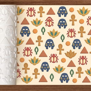 Può includere: Un mattarello cilindrico bianco con un motivo in rilievo di simboli egizi, accanto a un foglio di pasta beige chiaro con una stampa ripetuta di scarabei rossi, maschere di faraone blu, fiori di loto gialli, ankh marroni e triangoli.