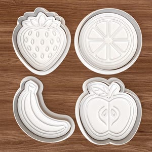 Puede incluir: Cuatro cortadores de galletas de plástico blanco con forma de fresa, rodaja de naranja, plátano y manzana. Los cortadores están sobre una superficie de madera. La fresa tiene semillas detalladas y la manzana muestra el corazón. La rodaja de naranja tiene detalles segmentados.