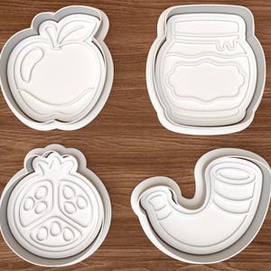 Op de afbeelding: Een set van vier witte plastic koekjesvormen met verschillende vormen: een appel met een blaadje, een glazen pot met een etiket, een dwarsdoorsnede van een granaatappel met zaden en een gebogen hoornvormig object met ribbels.