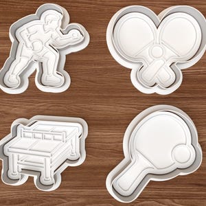 Può includere: Un set di quattro tagliabiscotti in plastica bianca a tema tennis da tavolo. Le forme includono una persona che gioca a ping pong, due racchette incrociate con una pallina, un tavolo da ping pong e una singola racchetta con una pallina.