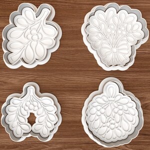 Op de afbeelding: Vier witte plastic koekjesvormers met ingewikkelde botanische ontwerpen. De vormen omvatten een maretak, een bloemenboeket met een strik, een arrangement van bladeren en een rond ornament, allemaal voorzien van gedetailleerde reliëfafdrukken.
