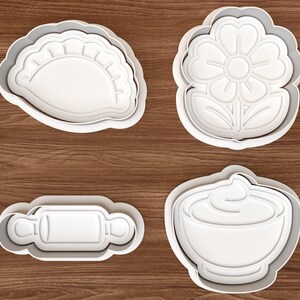 Puede incluir: Un juego de cuatro cortadores de galletas de plástico blanco que incluye una forma de empanadilla en media luna, una flor de seis pétalos con hojas, un rodillo de cocina y un cuenco con un remolino de crema, con detalles en relieve.