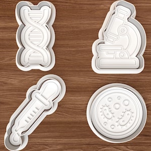 May include: Four white plastic cookie cutters in the shapes of science-related objects. The shapes include a DNA strand, a microscope, a pipette, and a petri dish with bacteria.