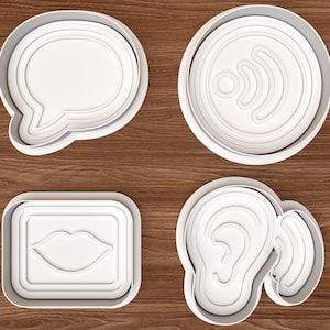 Puede incluir: Un juego de cuatro cortadores de galletas de plástico blanco con formas distintas: un globo de texto, un símbolo de Wi-Fi circular, un marco rectangular con labios en el centro y una forma de oreja. Cada cortador tiene un borde elevado.