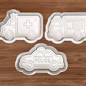 Peut inclure: Trois emporte-pièces en plastique blanc en forme de véhicules d'urgence. L'ensemble comprend une ambulance avec une croix, un camion de pompiers et une voiture de police avec le mot « POLICE ». Les emporte-pièces sont sur une surface en bois.