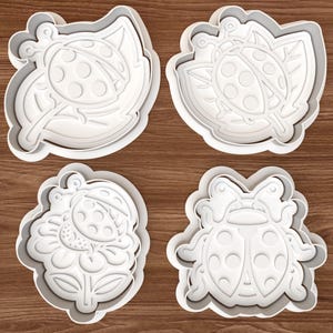 May include: Four white plastic cookie cutters in the shape of ladybugs and leaves. The cutters are arranged in a square, each with a different design. The ladybugs have detailed wing and body patterns.