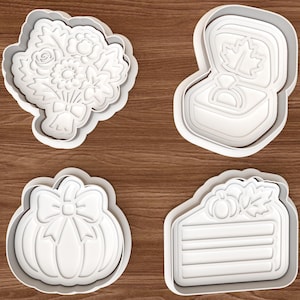 May include: A set of four white plastic cookie cutters featuring distinct shapes: a floral bouquet, an open ring box with a ring inside, a pumpkin adorned with a bow, and a slice of cake topped with a small pumpkin and a leaf design.