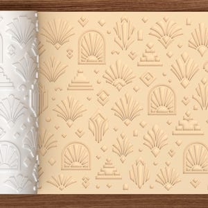 May include: A white cylindrical rolling pin with a repeating embossed pattern of sunburst arches, stepped pyramids, and geometric diamonds. The pattern is shown imprinted into a light beige dough surface, creating a textured relief design.
