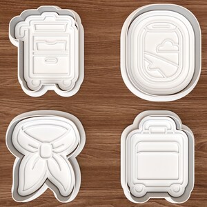 以下が含まれることがあります： 旅行をテーマにした4種類の白いプラスチック製クッキー型セット。機内サービス用カート、雲と翼が見える楕円形の飛行機の窓、ネックスカーフ、ハンドルと車輪が付いたキャリーケースの形をしています。