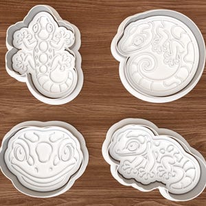 Puede incluir: Un juego de cuatro cortadores de galletas de plástico blanco con diseños en relieve detallados de varios lagartos y geckos. Cada cortador tiene una forma distinta, incluyendo un gecko de cuerpo entero, un lagarto enroscado, una cara de gecko y un lagarto en reposo.