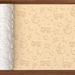May include: A white cylindrical rolling pin with a repeating embossed pattern of roller skates, disco balls, hearts, stars, and lightning bolts. The rolling pin is shown next to a light tan surface displaying the same pattern imprinted in relief.