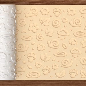May include: A white cylindrical rolling pin with a raised pattern of snails, flowers, mushrooms, clouds, leaves, and hearts. The rolling pin is shown next to a light beige sheet of dough that has been embossed with the same repeating nature-themed design.