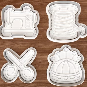 Può includere: Quattro formine per biscotti in plastica bianca a forma di oggetti legati al cucito. Le forme includono una macchina da cucire, una bobina di filo, forbici e un cuscinetto per spilli con un dettaglio a cuore. Le formine sono su una superficie di legno marrone.