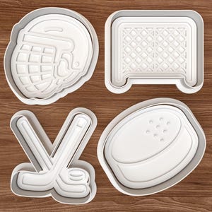 May include: Four white plastic cookie cutters in the shape of hockey equipment. The shapes include a hockey helmet, a hockey net, crossed hockey sticks, and a hockey puck. The cutters are on a wooden surface.