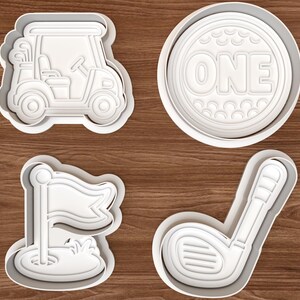 May include: A set of four white plastic cookie cutters featuring golf-themed designs: a golf cart, a circular cutter with the text ONE inside a golf ball pattern, a golf flag on a putting green, and a golf club with a textured grip and head.