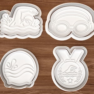 Könnte beinhalten: Vier weiße Ausstecher aus Kunststoff in Schwimm-Themen-Formen. Designs umfassen einen Schwimmer, Schwimmbrille, Wasserspritzer und eine Medaille mit einem Schwimmer und Lorbeerkranz. Die Ausstecher befinden sich auf einer Holzmaserungsoberfläche.