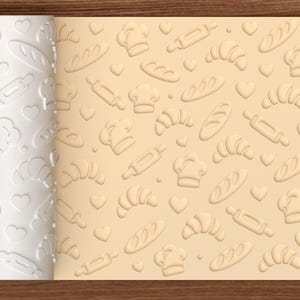 May include: A white cylindrical rolling pin with an embossed pattern of chef hats, rolling pins, croissants, baguettes, and hearts. The rolling pin is shown next to a light tan dough sheet featuring the same embossed baking-themed design.