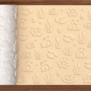 May include: A white cylindrical rolling pin with a repeating embossed pattern of frogs, water lilies, cattails, snails, and water droplets, shown alongside a light beige flat surface displaying the same impressed pond-themed design.