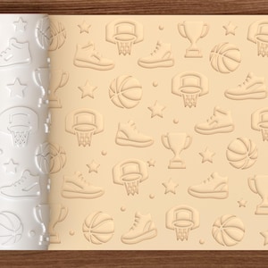 May include: A cylindrical rolling pin with a white surface featuring an embossed pattern of basketballs, sneakers, trophies, basketball hoops, and stars, shown alongside a flat sheet of light tan dough imprinted with the same repeating design.