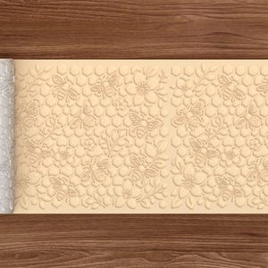 Può includere: Un mattarello bianco con un motivo a nido d'ape e api viene srotolato su una superficie beige con lo stesso disegno. Il disegno comprende api, fiori e forme a nido d'ape. Lo sfondo è una superficie di legno.