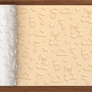 Puede incluir: Un rodillo cilíndrico blanco con un patrón en relieve repetido de ositos de peluche, biberones, sonajeros, nubes, estrellas y círculos pequeños, junto a una lámina de masa color beige claro con el mismo diseño impreso.