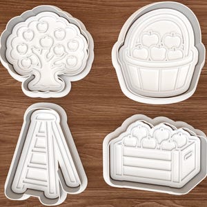 Puede incluir: Un juego de cuatro cortadores de galletas de plástico blanco que representan un manzano, una cesta llena de manzanas, una escalera alta y una caja de madera con varias manzanas. Cada cortador tiene un contorno y detalles en relieve.