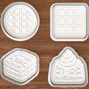 Op de afbeelding: Een set van vier witte plastic koekjesvormen met wafelthema. De vormen bestaan uit een ronde wafel, een vierkante wafel, een stapel wafels met een klontje boter en een enkele wafel met een toef slagroom of siroop erop.
