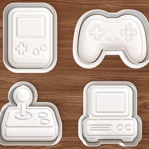 Peut inclure: Un ensemble de quatre emporte-pièces en plastique blanc en forme d'équipement de jeu rétro, comprenant une console portable, une manette, un joystick avec socle et une console d'ordinateur de bureau, avec des détails intérieurs en relief.