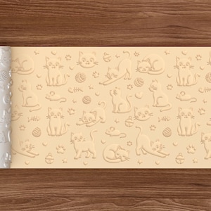 May include: A white rolling pin with a repeating embossed pattern of cats, fish bones, paw prints, and yarn balls. The pattern is displayed on a light tan surface. The rolling pin is partially rolled out, revealing the pattern.