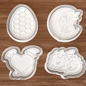Puede incluir: Cuatro cortadores de galletas de plástico blanco con formas de dragón. Las formas incluyen un huevo con un patrón de escamas, un dragón en una luna creciente, un corazón con alas de dragón y un bebé dragón durmiente. Los cortadores están sobre una superficie de madera.