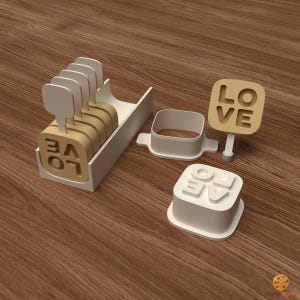 May include: A set of white plastic cookie cutters and stamps featuring the word LOVE in a square frame. The set includes a rectangular storage tray holding several stamps, a square cutter frame, and a stamp with a handle displaying the word LOVE.