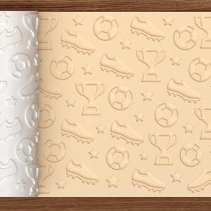 May include: A cylindrical rolling pin with a white surface featuring an embossed pattern of soccer balls, cleats, trophies, and stars. The rolling pin is shown next to a light tan flat surface displaying the same pattern imprinted in relief.