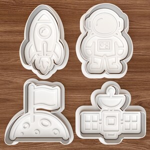 May include: Four white plastic cookie cutters in space-themed shapes: a rocket, an astronaut, a moon with a flag, and a satellite. The cutters are on a brown wooden surface.