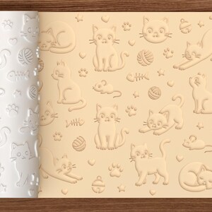 May include: A white cylindrical rolling pin with an embossed pattern of cats, yarn balls, fish bones, mice, bells, hearts, and paw prints. The pattern is shown imprinted on a light beige dough surface, featuring repeating feline-themed motifs.