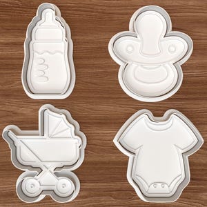 Puede incluir: Cuatro cortadores de galletas de plástico blanco con formas de bebé: un biberón, un chupete, un cochecito y un body. Los cortadores están sobre una superficie de madera marrón. Los cortadores de galletas son para hornear.