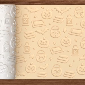 May include: A white cylindrical rolling pin with a repeating embossed pattern of jack-o'-lanterns, bats, lanterns, and candy buckets, shown alongside a light beige dough sheet displaying the same impressed Halloween-themed design.