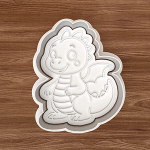 May include: A white plastic dragon-shaped cookie cutter. The dragon has a friendly face, wings, and a long tail. The cutter is surrounded by a gray border and sits on a wooden surface. The cookie cutter is approximately 4 inches in length.
