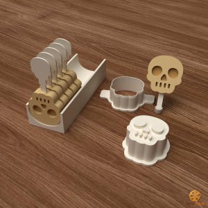 May include: A set of beige skull-shaped cookie cutters and matching sticks, including a white storage tray, a separate skull cutter, and a skull-shaped stamp. The items are arranged to show the components for creating skull-shaped treats on sticks.