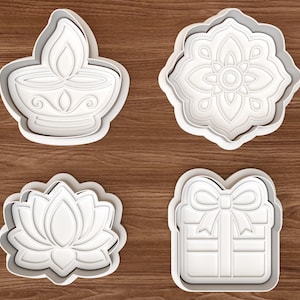 May include: A set of four white plastic biscuit cutters featuring distinct shapes: a diya oil lamp, a symmetrical floral mandala, a blooming lotus flower, and a square gift box tied with a decorative bow, all displayed on a wood grain surface.