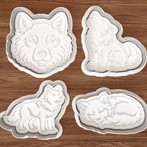 Puede incluir: Cuatro cortadores de galletas de plástico blanco con forma de lobos. Uno es una cabeza de lobo, otro un lobo aullando, un tercero un lobo de pie y el último un lobo durmiendo. Los cortadores están sobre una superficie de madera marrón.