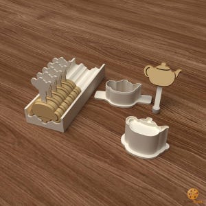 Puede incluir: Una bandeja de plástico blanco que contiene varios palitos de madera de color marrón claro con forma de tetera, junto a dos cortadores de galletas de plástico blanco con forma de tetera y un palito de madera sobre una base.
