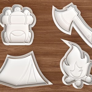 May include: Four white plastic cookie cutters in camping shapes: a backpack, an axe, a tent, and a campfire. The cutters are on a wooden surface. The cookie cutters are for making camping-themed cookies.