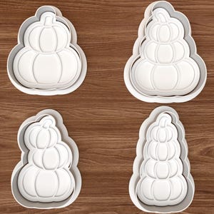 May include: Four white plastic cookie cutters in the shapes of stacked pumpkins. The top left cutter features two pumpkins, the top right features three, the bottom left features three in a staggered arrangement, and the bottom right features four.